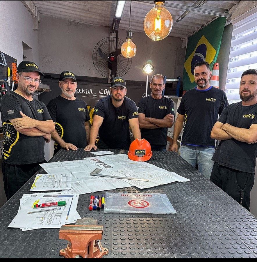 Equipe da Volt's Soluções em Elétrica reunida na oficina, com projetos elétricos sobre a bancada e capacete da empresa