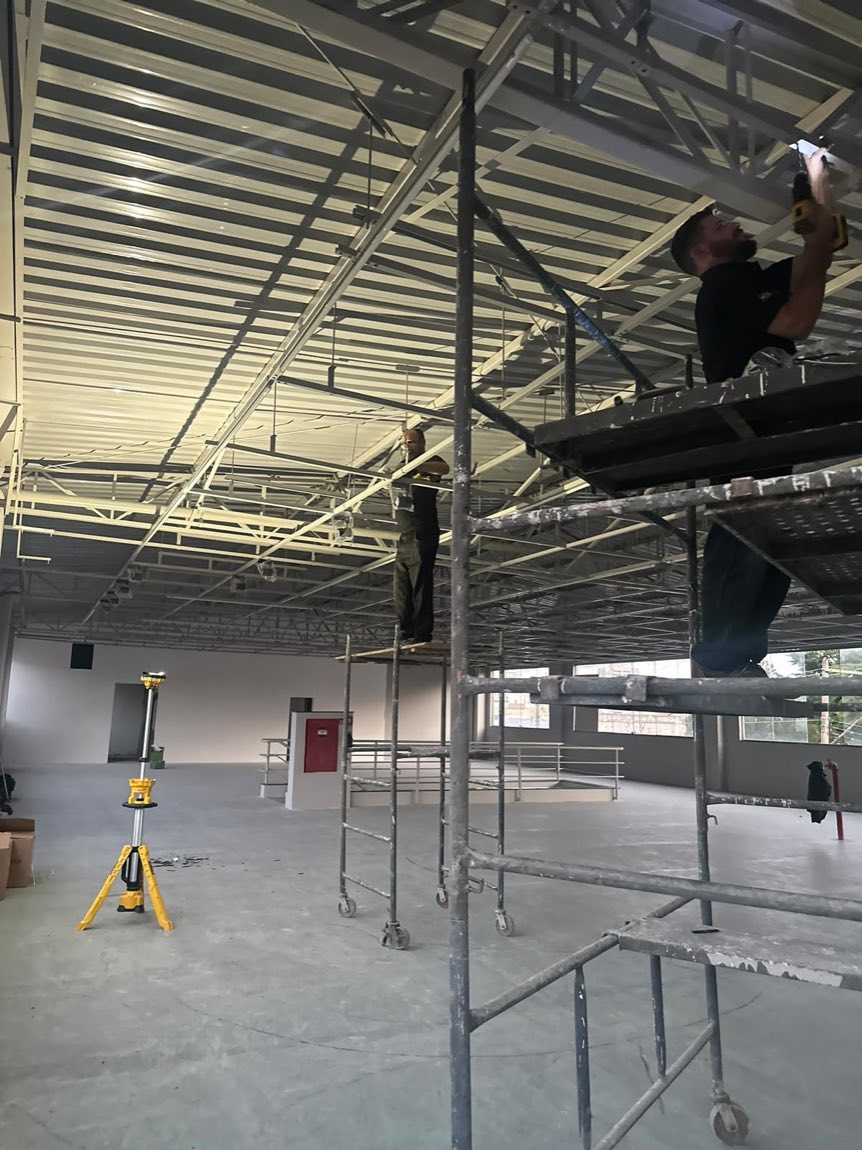 Dois profissionais em andaimes instalando luminárias no forro metálico de um galpão